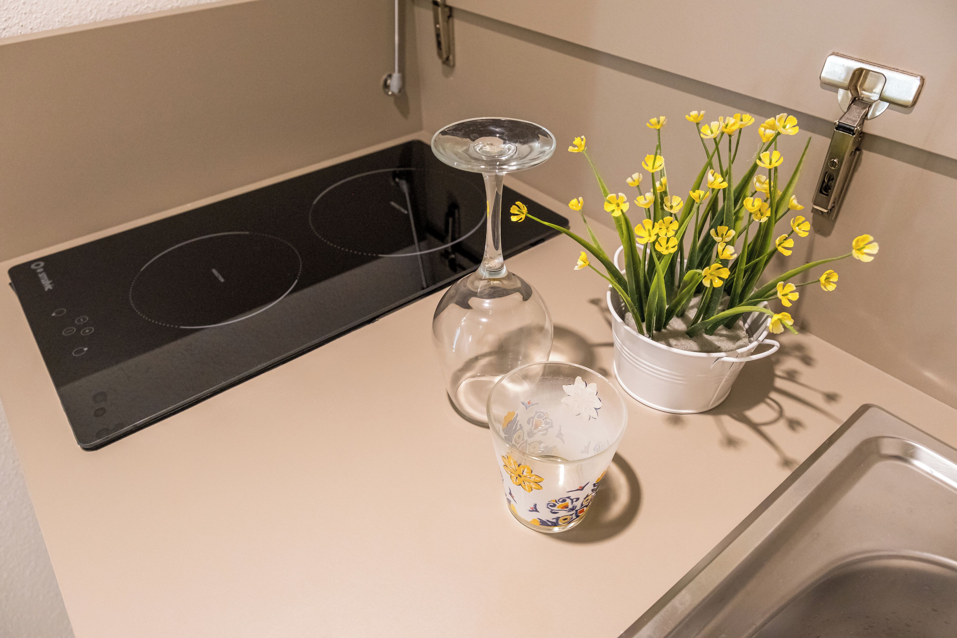 A kitchen with a black induction cooktop, a wine glass, and a potted plant with yellow flowers.
