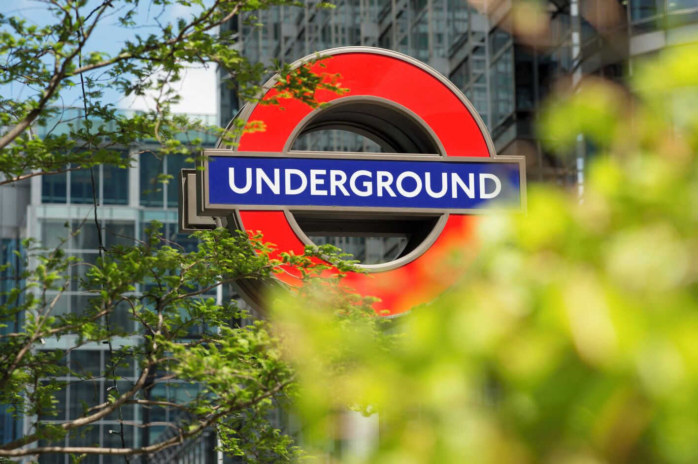 Indicador del metro de Londres amb la paraula 'UNDERGROUND' en blanc sobre fons blau, envoltat de verdura i edificis moderns.