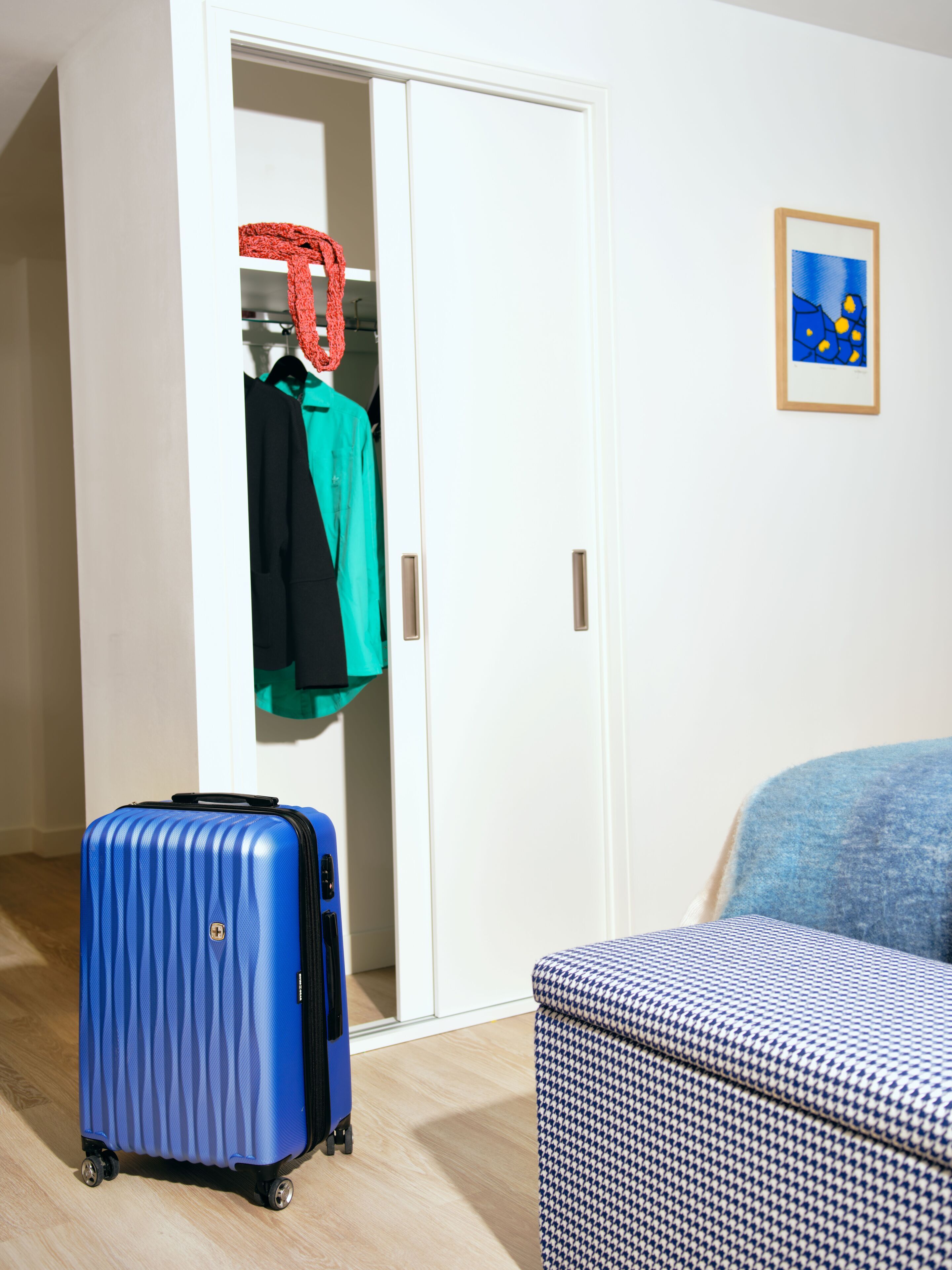 A room with a blue suitcase, a houndstooth-patterned chair, a wardrobe with clothes, and a framed abstract painting on the wall.
