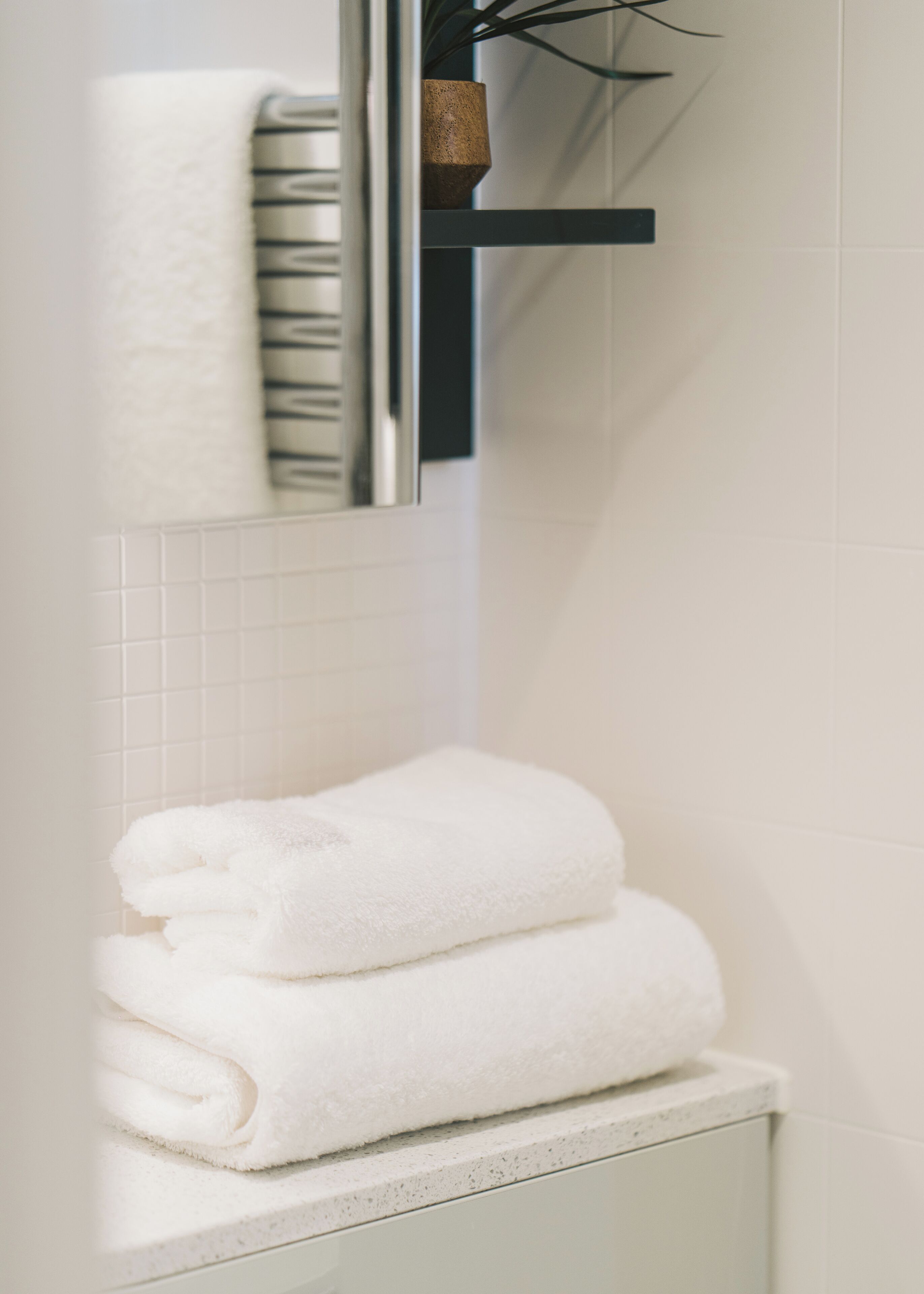 A bathroom with white tiles, a towel rack with folded towels, and a mirror reflecting the towel rack.