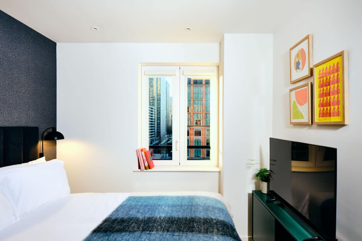 A modern hotel room with a large bed, a TV, a window with a city view, and artwork on the wall.