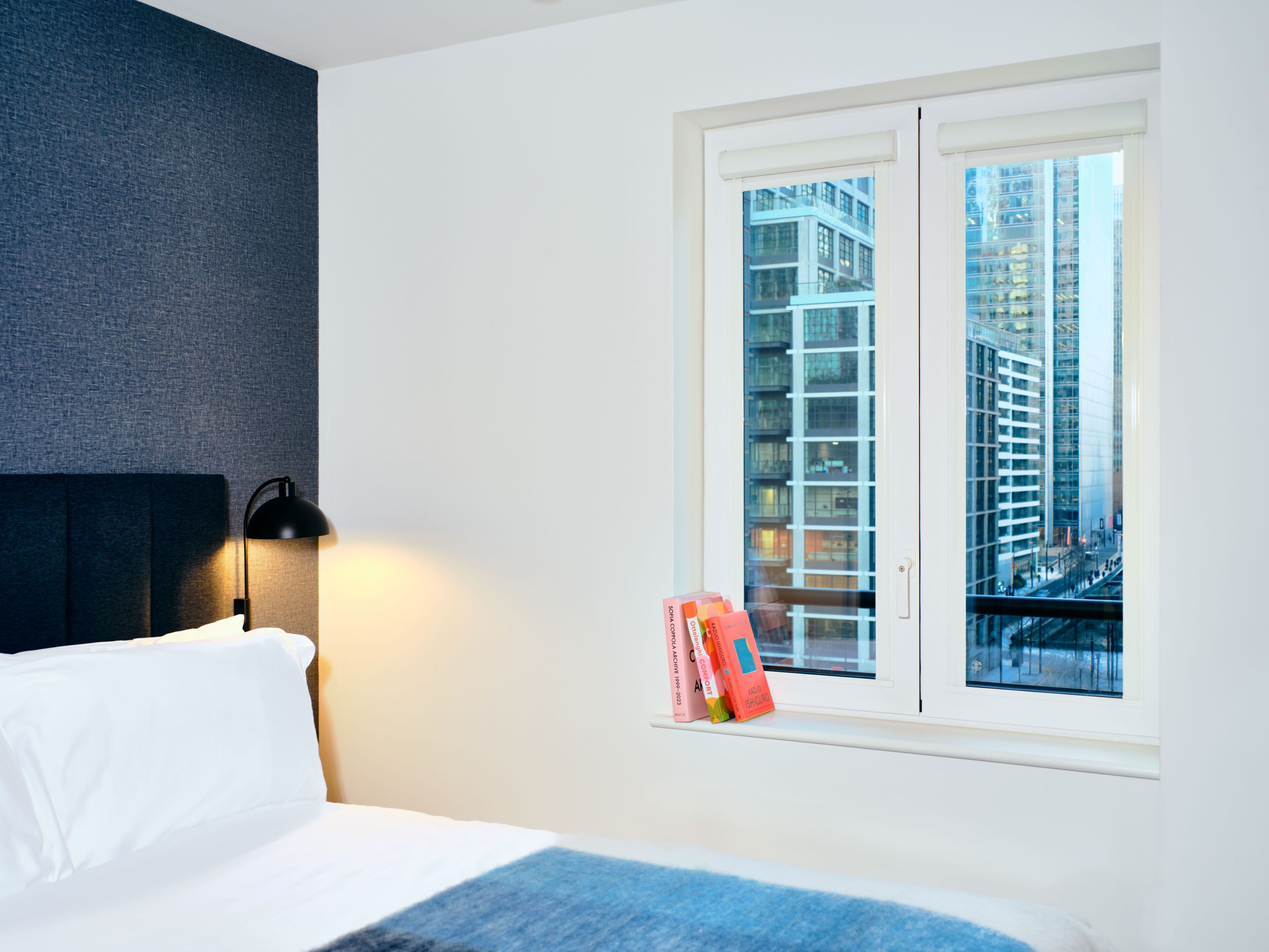 Una habitación de hotel con una cama, una mesita de noche, una lámpara, una ventana con vistas a edificios altos y algunos libros en el alféizar.