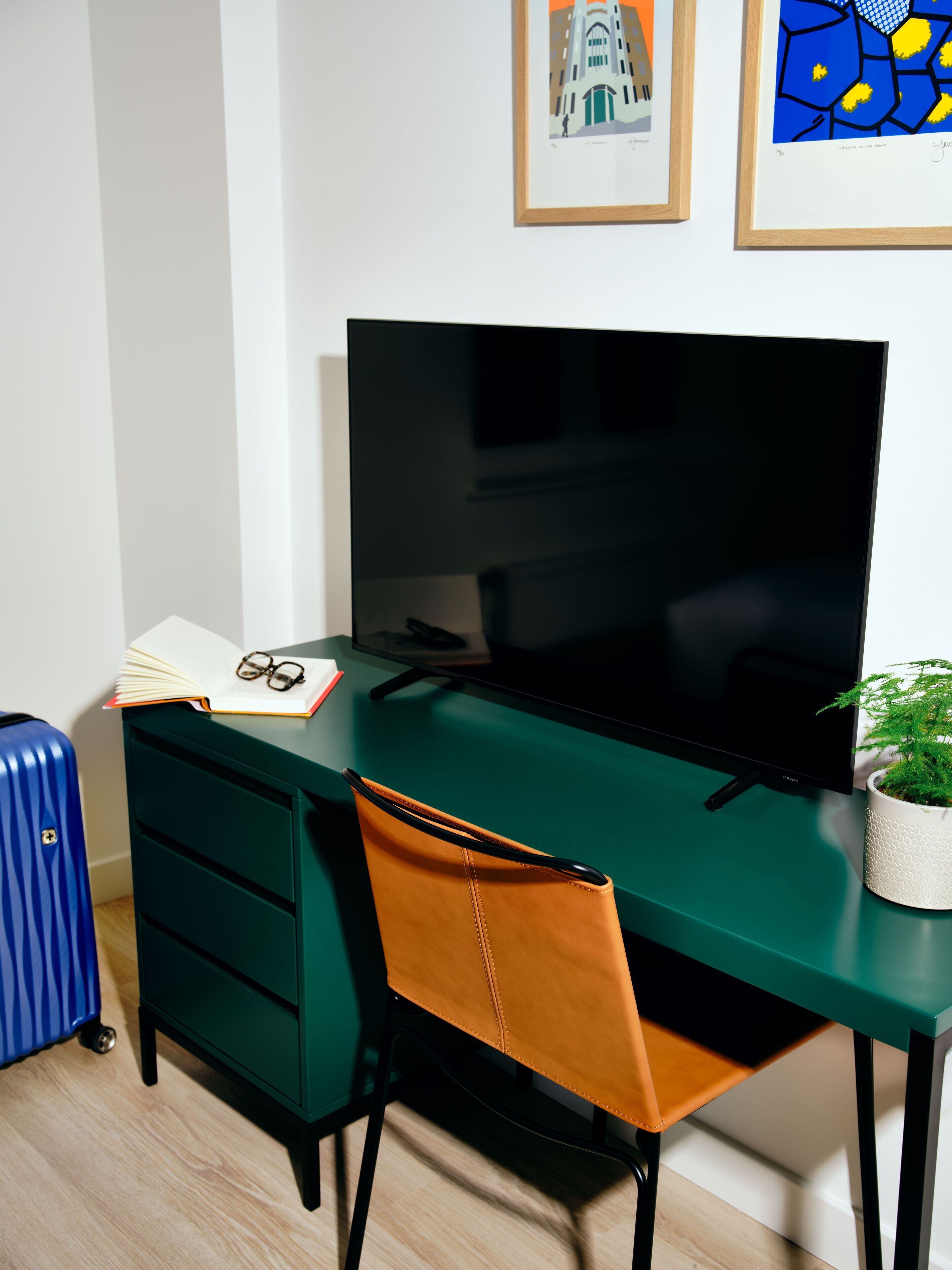 Una habitación moderna con un escritorio verde, un televisor negro, una maleta azul y un cuadro en la pared.