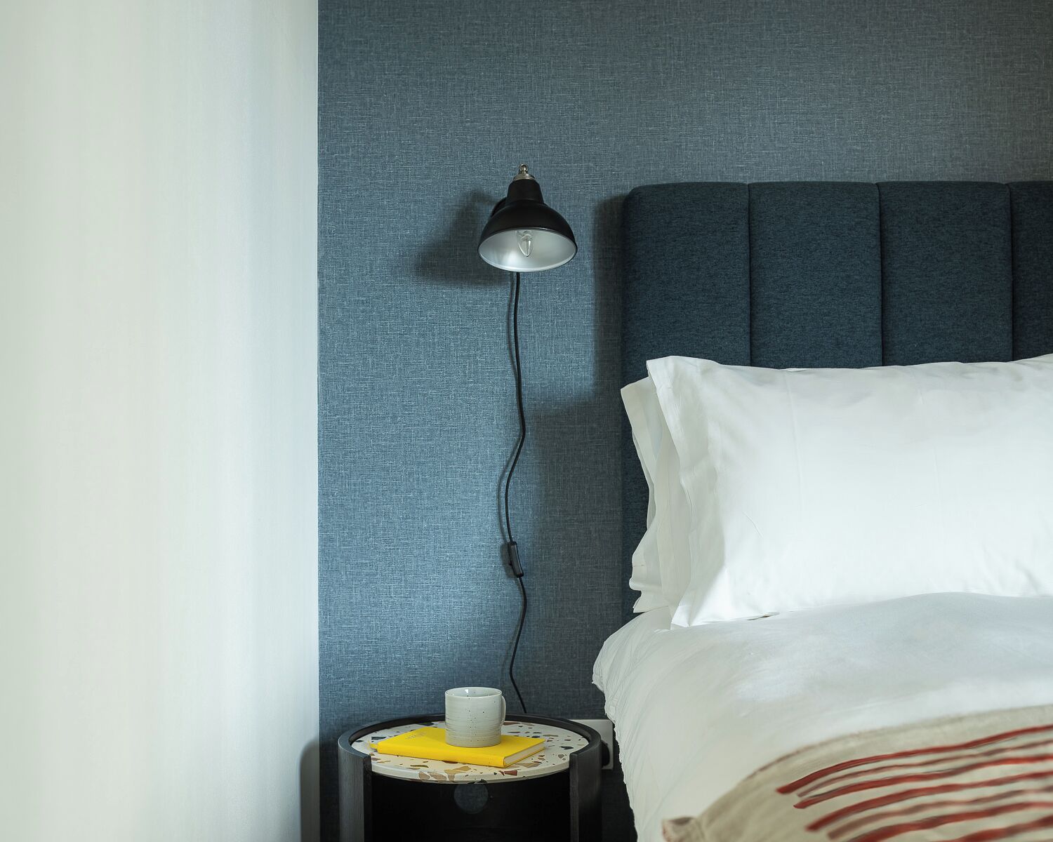 A neatly made bed with white sheets and a pillow, a bedside table with a lamp, and a wall with a textured pattern.