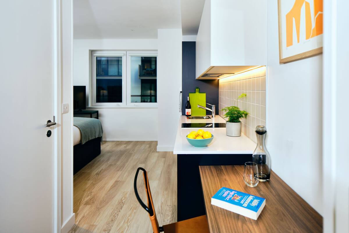 A modern kitchen with a wooden floor, a dining table with a bowl of lemons, a book, and a wine bottle.