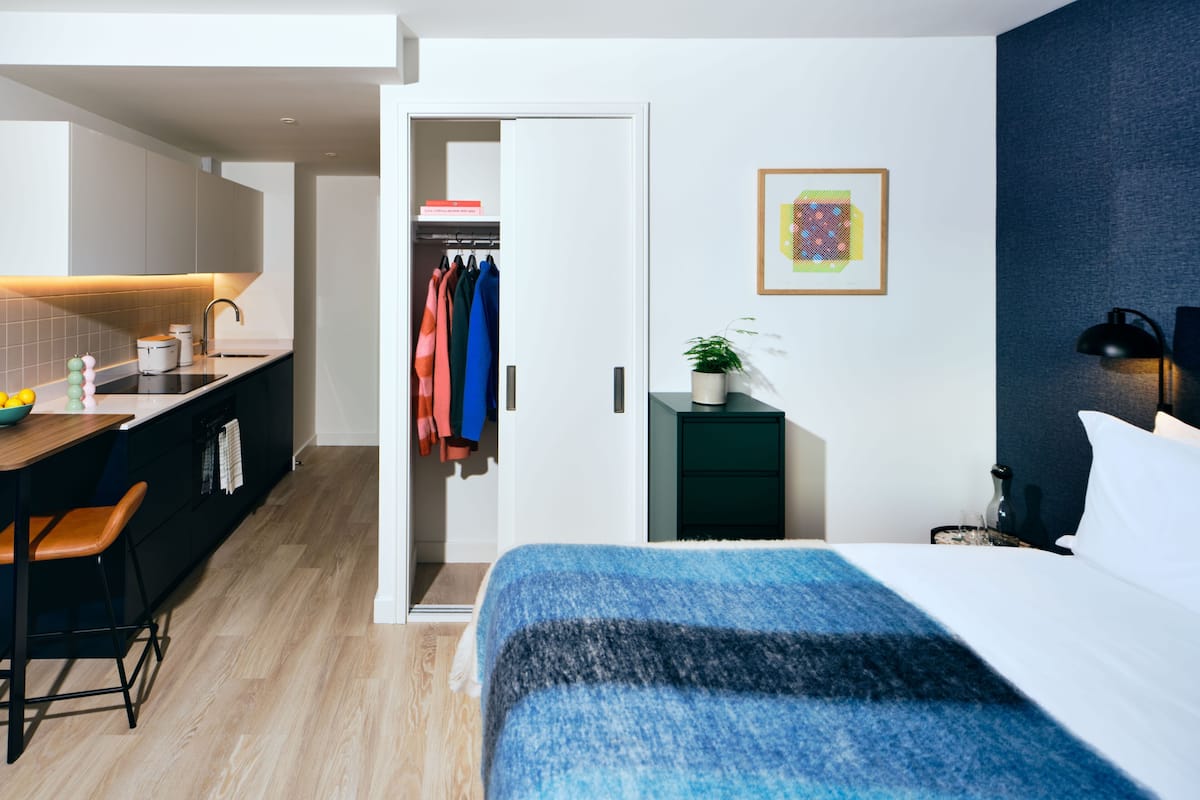A modern hotel room with a bed, a desk with a chair, a small side table with a lamp, a wardrobe, and a bathroom visible through an open door.