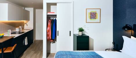 A modern hotel room with a bed, a desk with a chair, a small side table with a lamp, a wardrobe, and a bathroom visible through an open door.