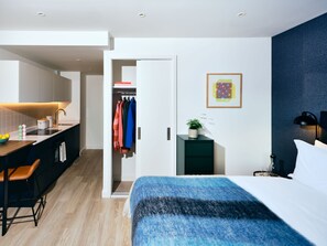 A modern hotel room with a bed, a desk with a chair, a small side table with a lamp, a wardrobe, and a bathroom visible through an open door. - Vertus Edit Canary Wharf (London)