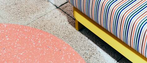 A modern outdoor seating area with a yellow chair and a colorful striped cushion, a terracotta planter with green plants, and a brick wall in the background.