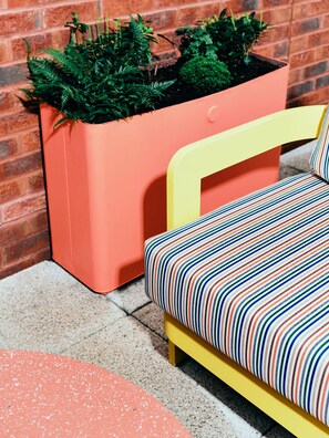 A modern outdoor seating area with a yellow chair and a colorful striped cushion, a terracotta planter with green plants, and a brick wall in the background. - Vertus Edit Canary Wharf (London)