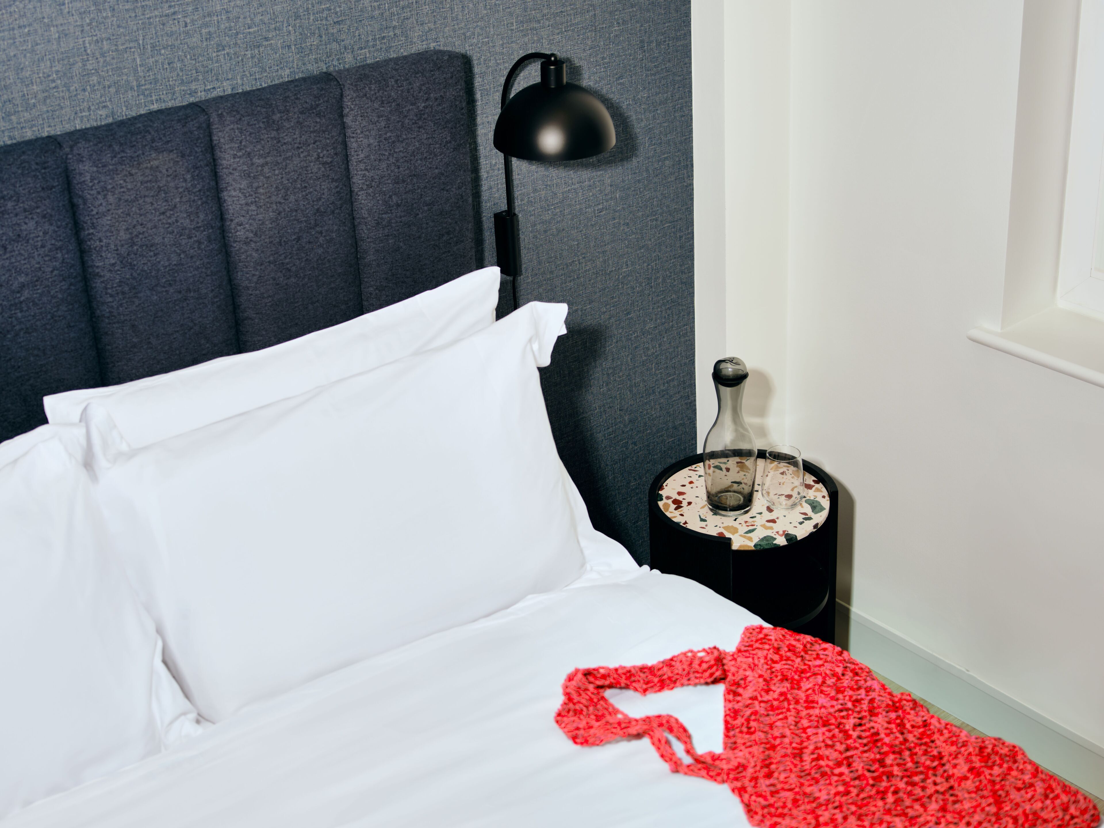 A neatly made bed with white linens, a grey headboard, a black lamp, and a small table with a bottle and glasses.