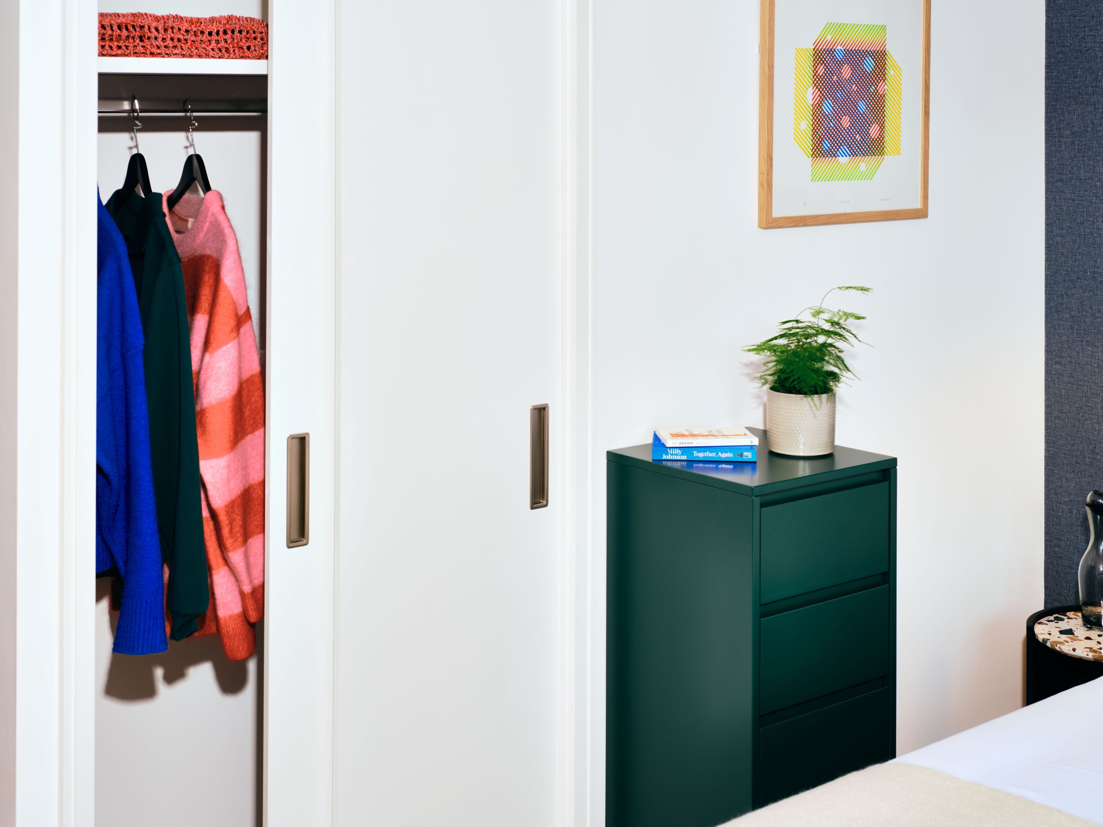A bedroom with a closet containing colorful clothing, a green dresser with a plant and a book, and a framed artwork on the wall.