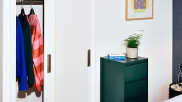 A bedroom with a closet containing colorful clothing, a green dresser with a plant and a book, and a framed artwork on the wall.