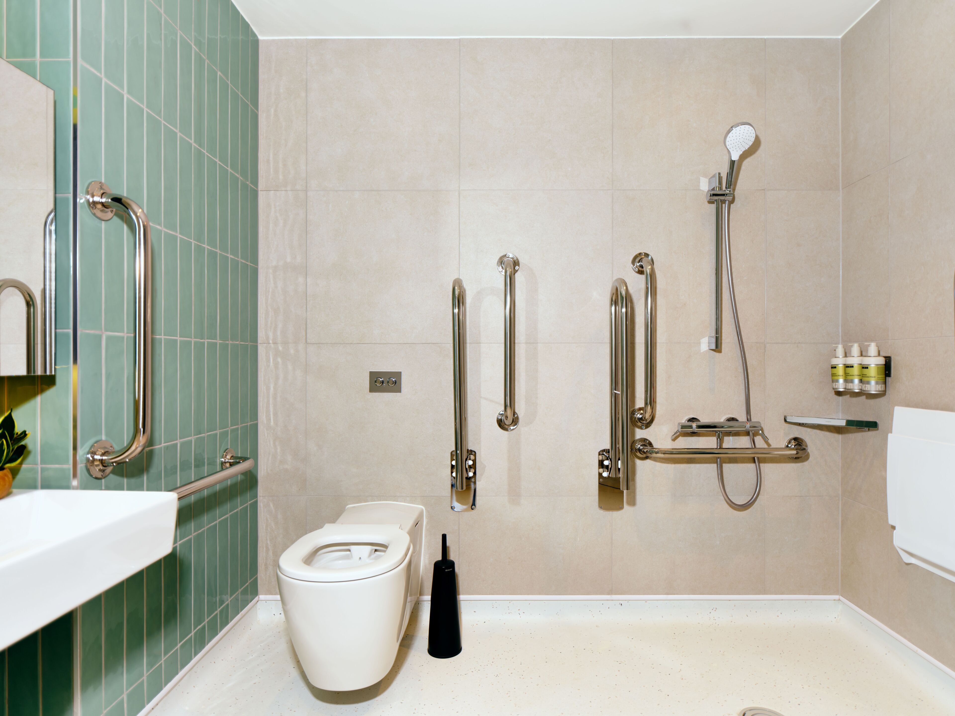 Ein Badezimmer mit Toilette, Haltegriffen, einer Dusche mit Duschbrause und einem Ablagebereich für Toilettenartikel.