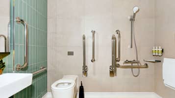 A bathroom with a toilet, grab bars, a shower with a handheld showerhead, and a shelf with toiletries.