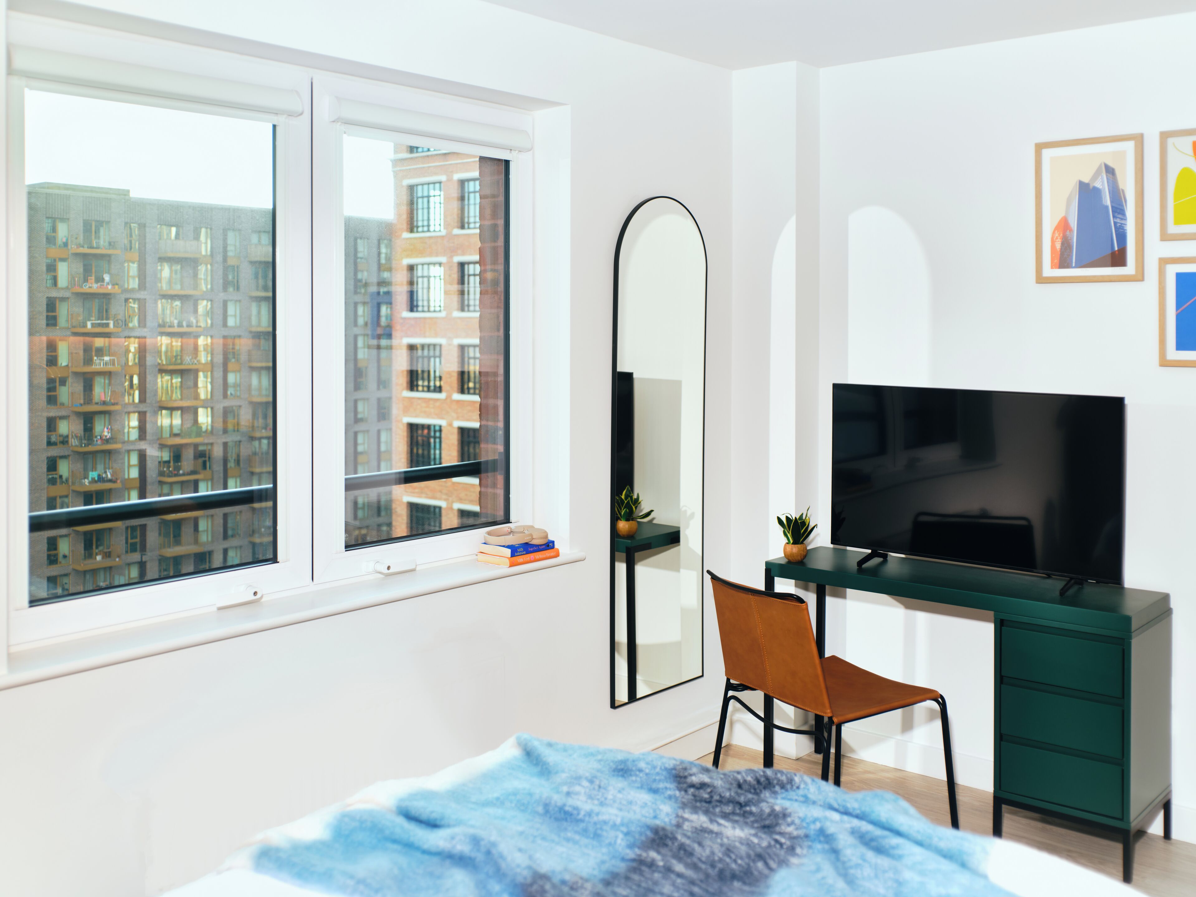 A modern bedroom with a large window, a desk with a TV, a chair, and a bed with a blue blanket.