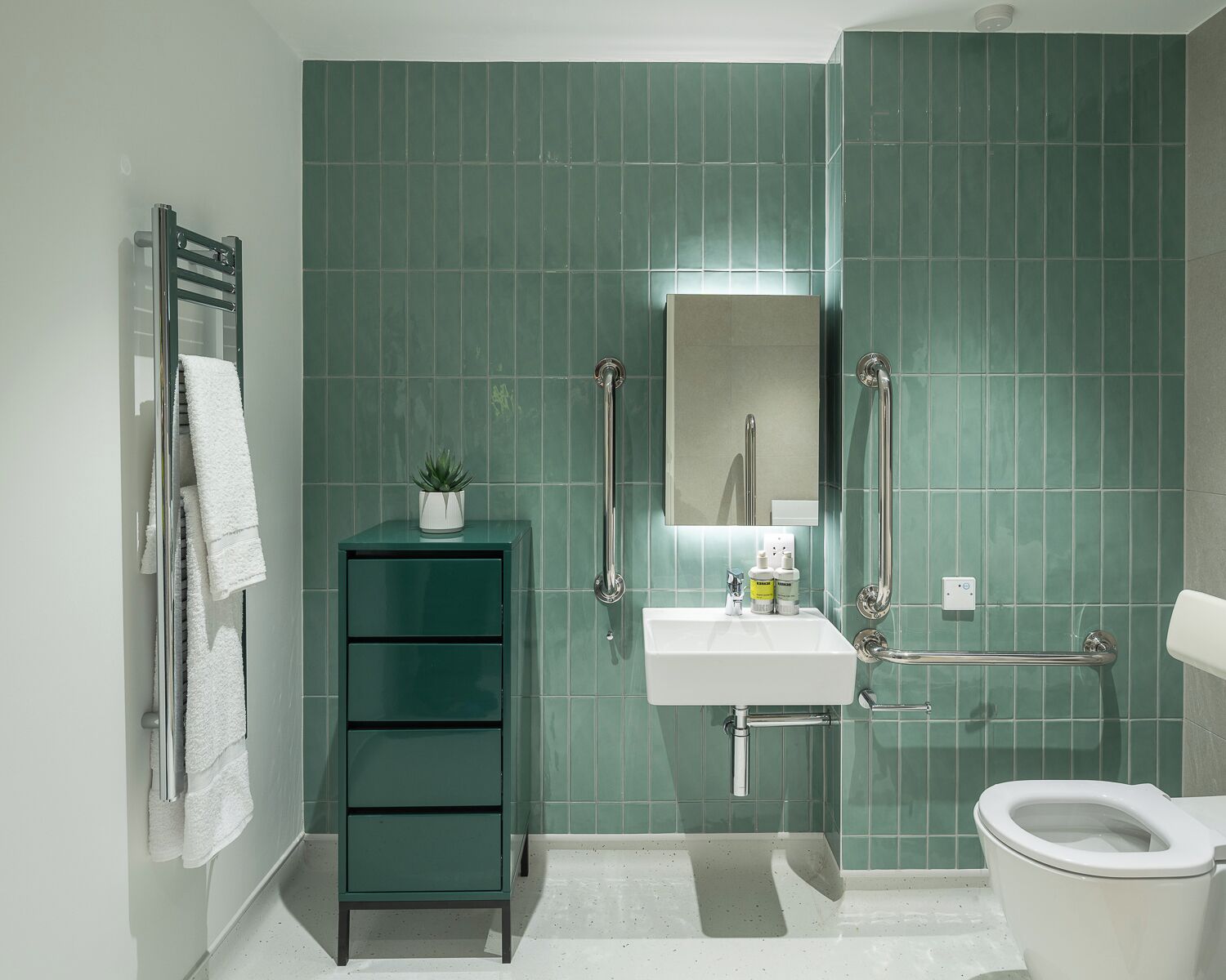 Un bagno con parete rivestita in piastrelle verdi, un lavandino bianco, uno specchio, un WC e un mobile contenitore con una pianta.