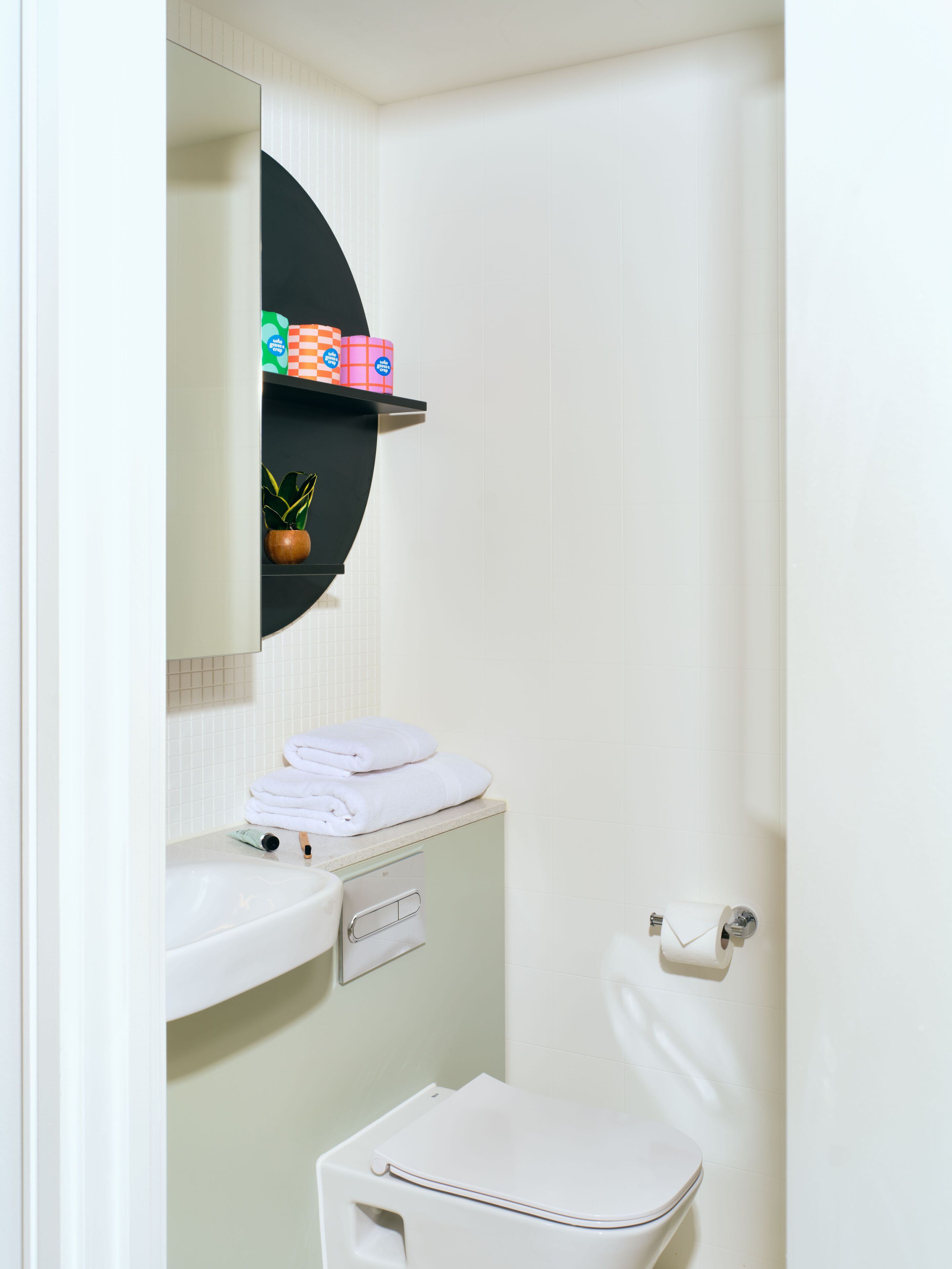 A bathroom with a white sink, a toilet, and a mirror above the sink. There are towels stacked on the sink and a potted plant on the shelf.
