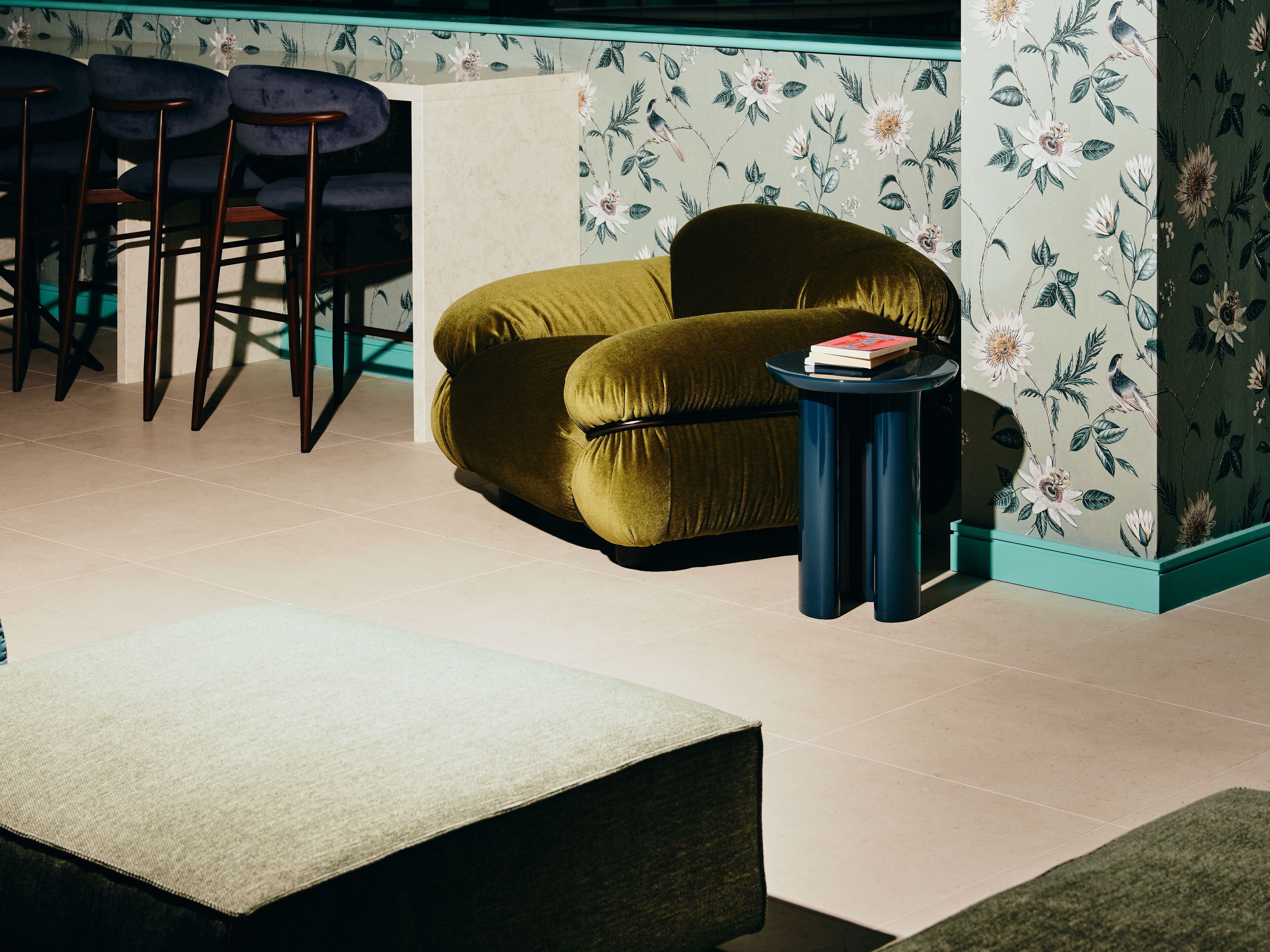 A room with a green sofa, a small round table with a book, and a floral wallpaper.