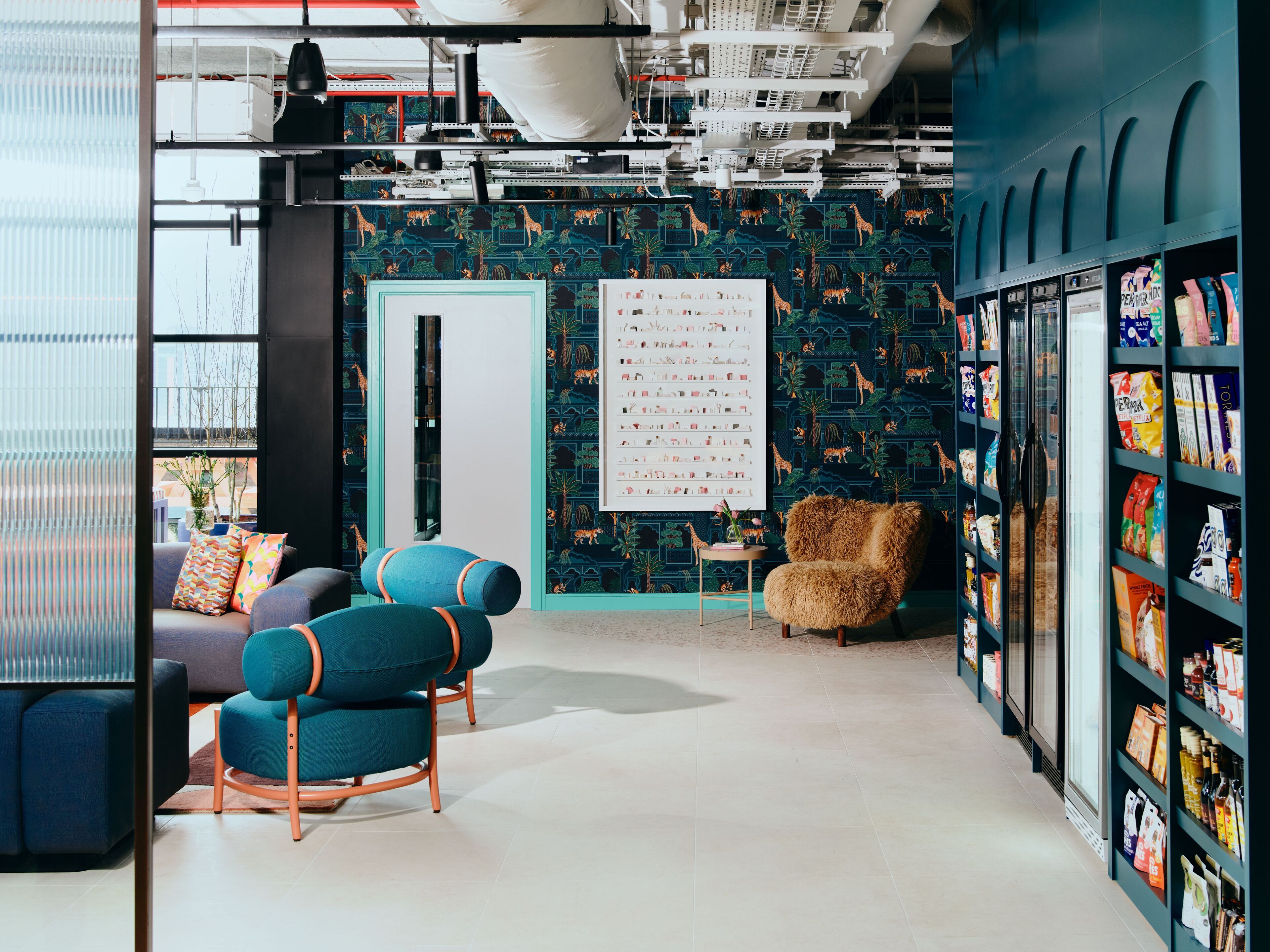 A modern interior with a teal patterned wall, a whiteboard, a seating area with blue chairs, and shelves with various items.