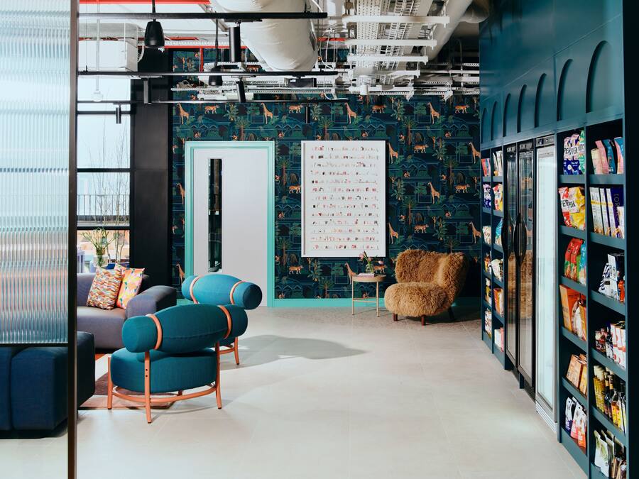 A modern interior with a teal patterned wall, a whiteboard, a seating area with blue chairs, and shelves with various items.
