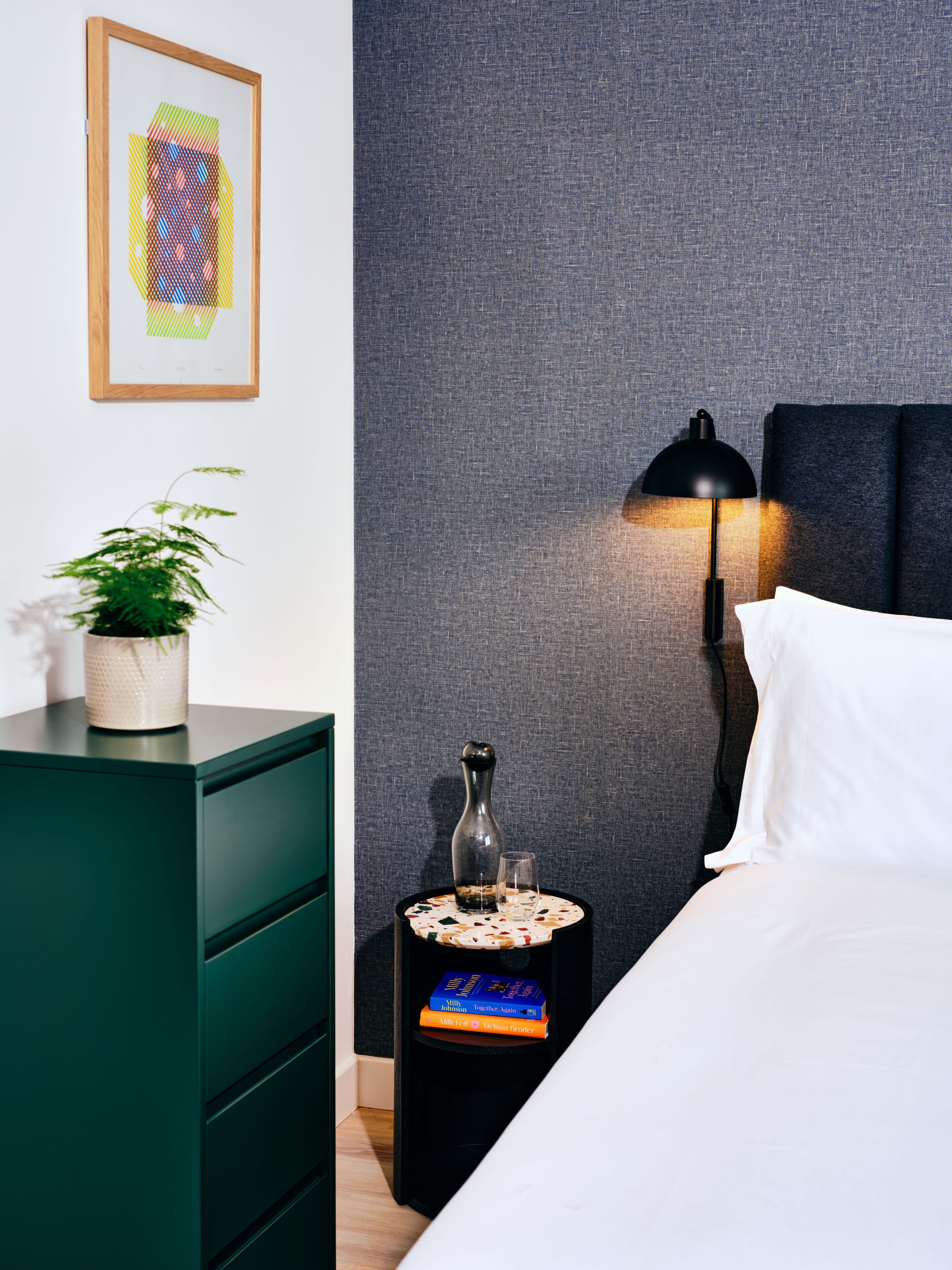 A neatly arranged bedroom with a bed, a bedside table with a lamp, a small side table with a vase and books, a green dresser, and a framed abstract artwork on the wall.