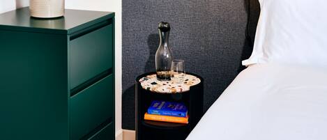 A neatly arranged bedroom with a bed, a bedside table with a lamp, a small side table with a vase and books, a green dresser, and a framed abstract artwork on the wall.