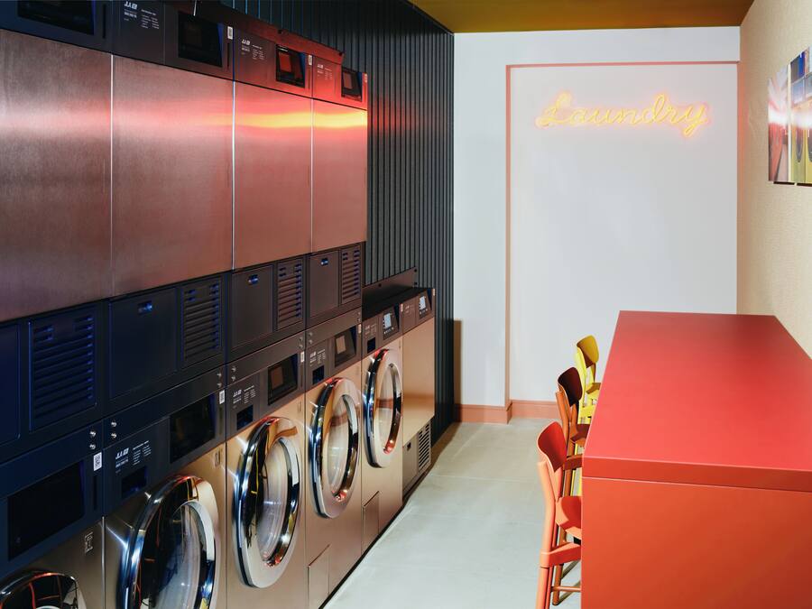 Een moderne wasserette met meerdere wasmachines en drogers, een neonbord met de tekst 'Laundry' en een rode balie met stoelen.