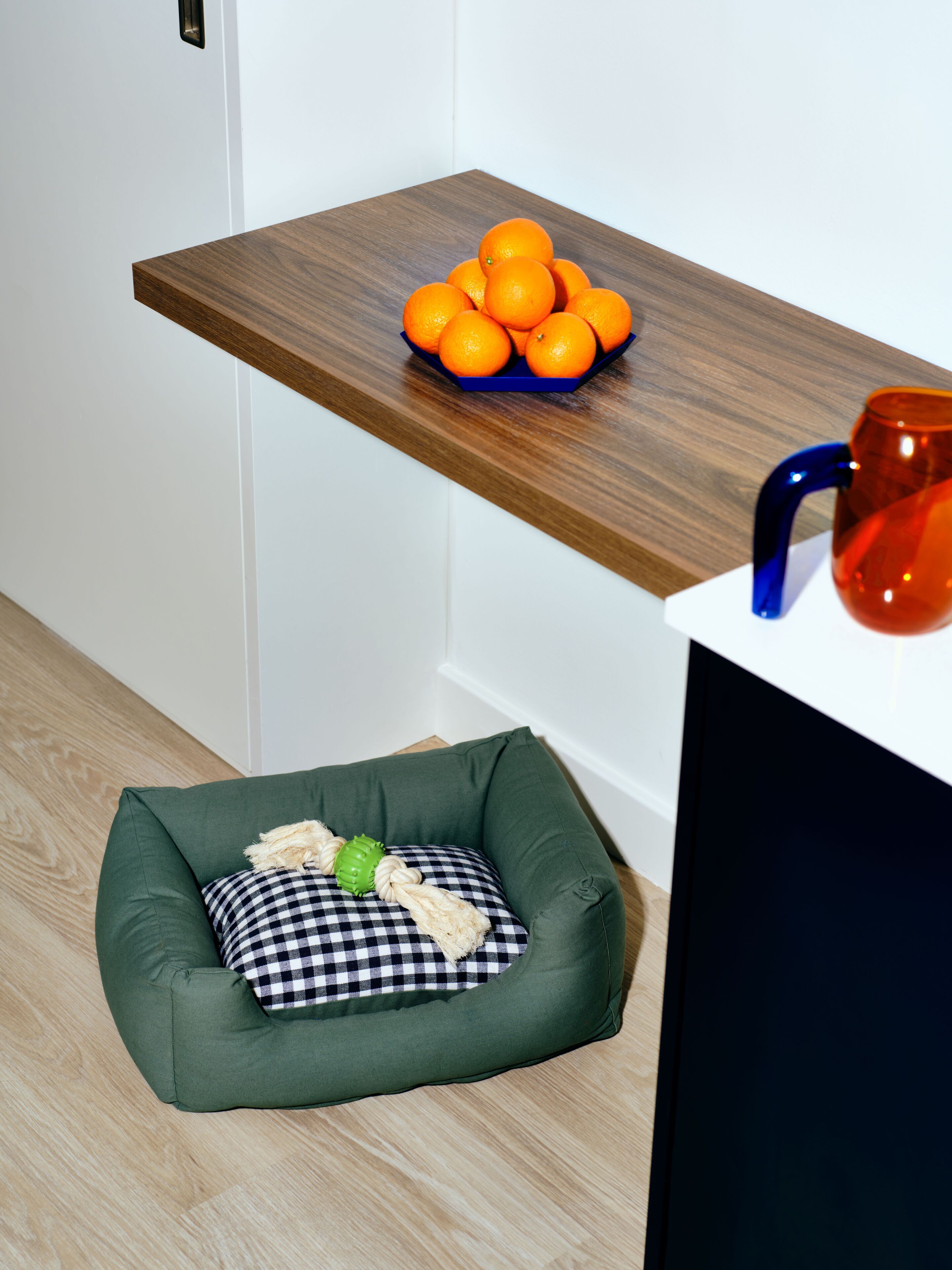 Una habitación con una estantería de madera con naranjas y una taza, una cama para mascotas verde con cojín a cuadros, y un armario negro.