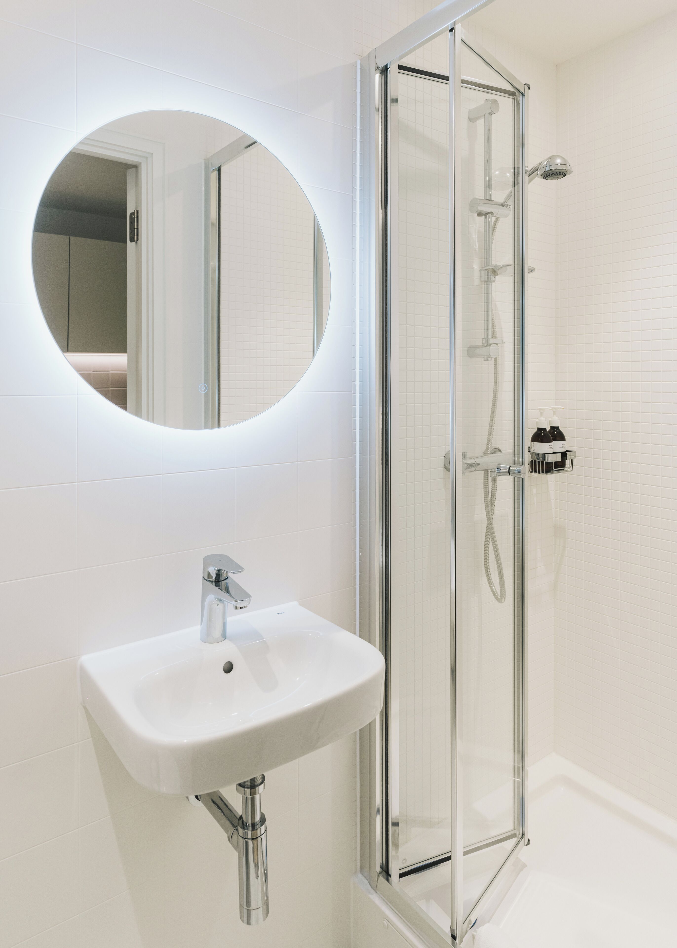 A bathroom with a glass-enclosed shower, a white sink, and a round mirror.