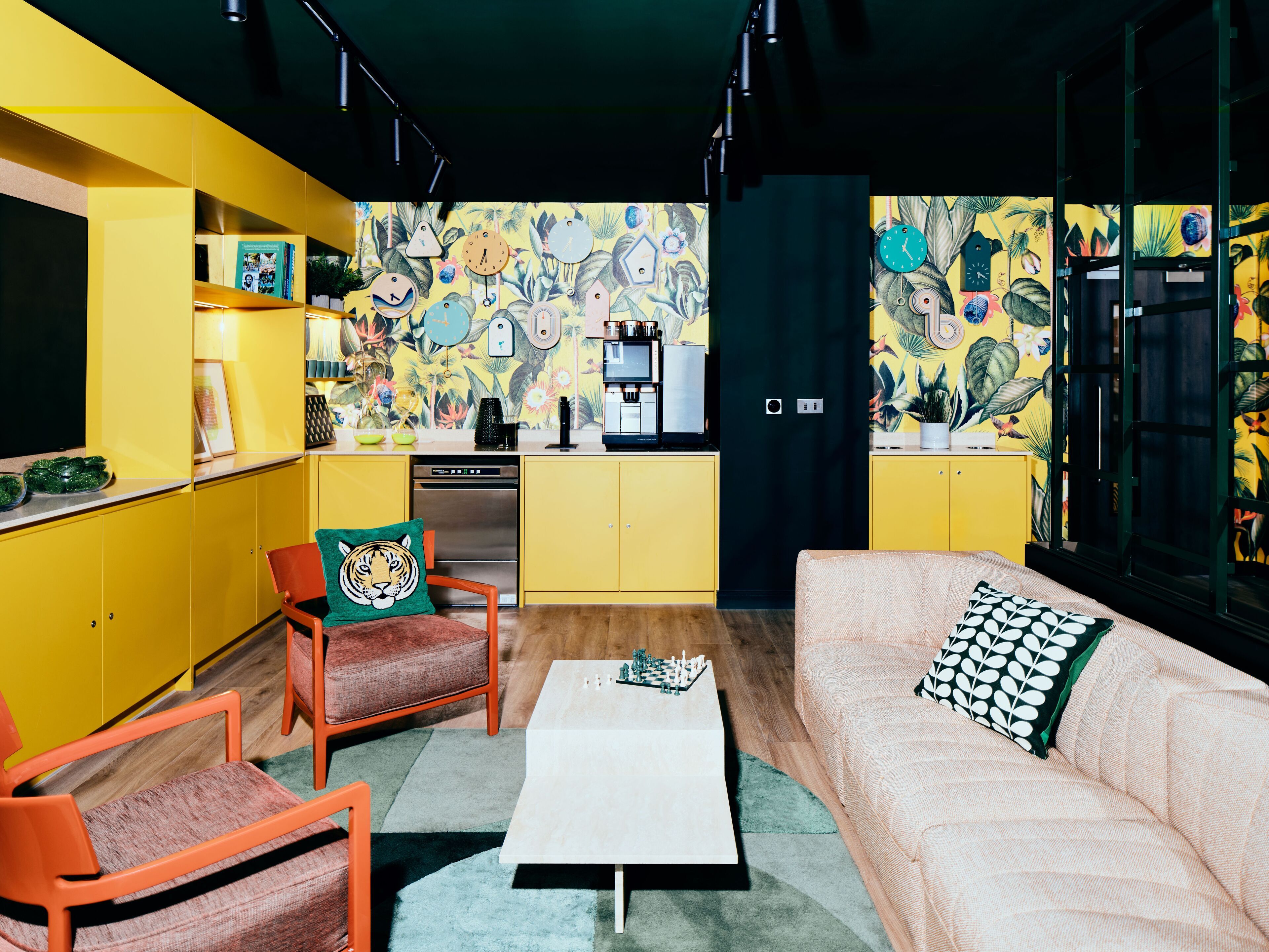 Une cuisine moderne avec des armoires jaunes, un réfrigérateur noir et une fresque murale colorée. On y trouve également un coin repas avec des chaises orange et une table basse blanche.