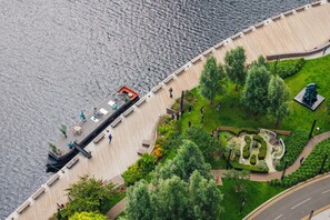 An aerial view of a waterfront promenade with a wooden walkway, benches, and landscaped gardens. - Vertus Edit Canary Wharf (London)