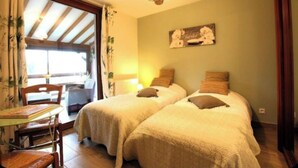 A room with two beds, a table, and a chair. There is a window with curtains and a light fixture above the beds. - Capucine's house (Les Moutiers-en-Cinglais)