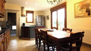 A dining area with a wooden table and chairs, a chandelier, and a kitchen in the background. - Capucine's house (Les Moutiers-en-Cinglais)