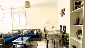 A living room with a sofa, coffee table, and dining table. There is a bookshelf with decorative items and a window with blinds. - Confort Moderne in the heart of Salé, near train station and airport (Rabat- Sale')