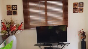 Ein Wohnzimmer mit Flachbildfernseher auf einem Schrank, Deckenventilator, einer Uhr und einem Fenster mit Jalousien.