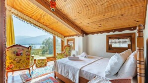 Ein Schlafzimmer mit Holzbalkendecke, einem Bett mit weißen Leinen, einem floralen Sessel und Blick auf einen See.