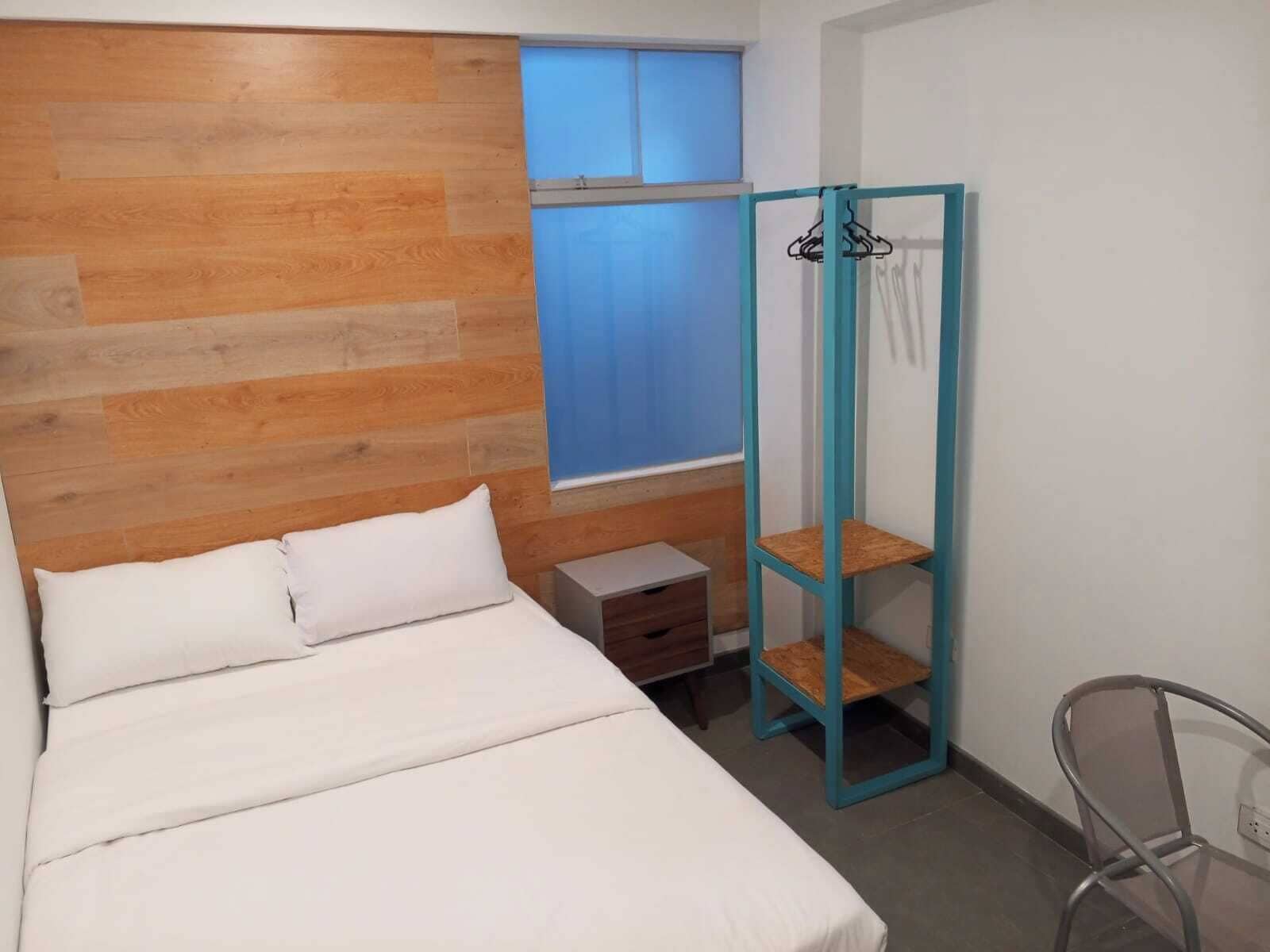 A bedroom with a wooden headboard, a bed with white bedding, a small bedside table, a window with blue curtains, and a blue wardrobe with a mirror.