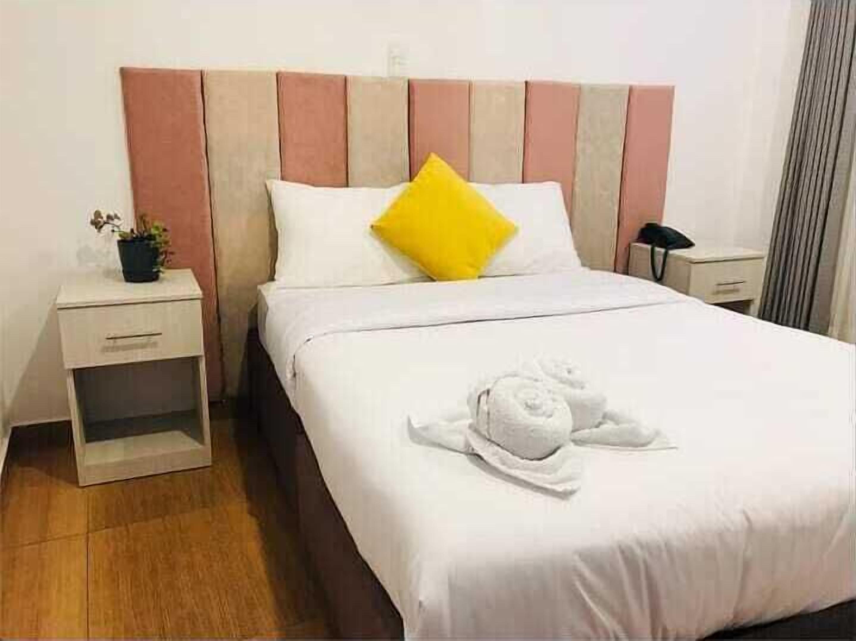 A neatly made bed with white bedding and a yellow pillow, a bedside table with a plant, and a small drawer unit.