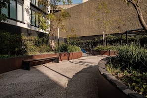 Stroll along the paved walkway to a serene seating area surrounded by lush greenery and a beautiful building with large windows.