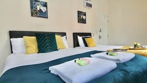A hotel room with two beds, each with a black headboard, white and green pillows, and a teal bedspread. There is a wooden side table with a tray of towels and toiletries. - LHR Studio 6 - Coventry (Coventry)