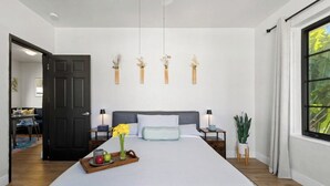 Un dormitori amb un llit gran, taules de nit, una petita taula auxiliar, una planta en test i una finestra amb cortines.