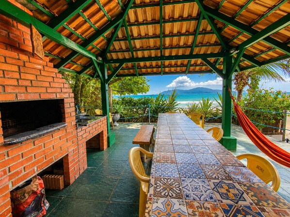 A covered outdoor seating area with a brick barbecue, a tiled table, and a view of the beach.