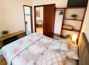 A bedroom with a bed, a wooden wardrobe, a bedside table with a lamp, and a small potted plant.