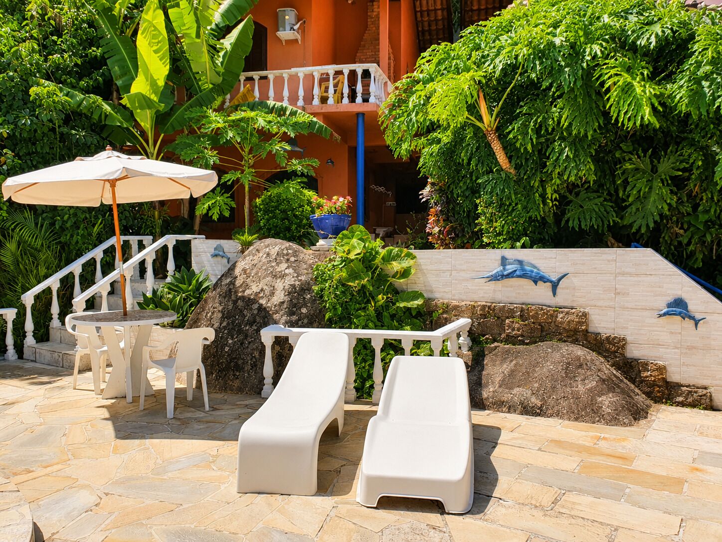 Un espace patio avec une table et des chaises blanches, un fauteuil de jardin blanc, un grand parasol et un mur en pierre orné de motifs de requins.