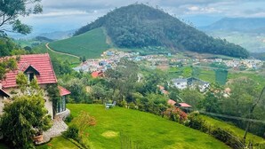 A hillside village with terraced fields, a prominent hill, and a few buildings with red roofs. - Stumpfields (Ootacamund)