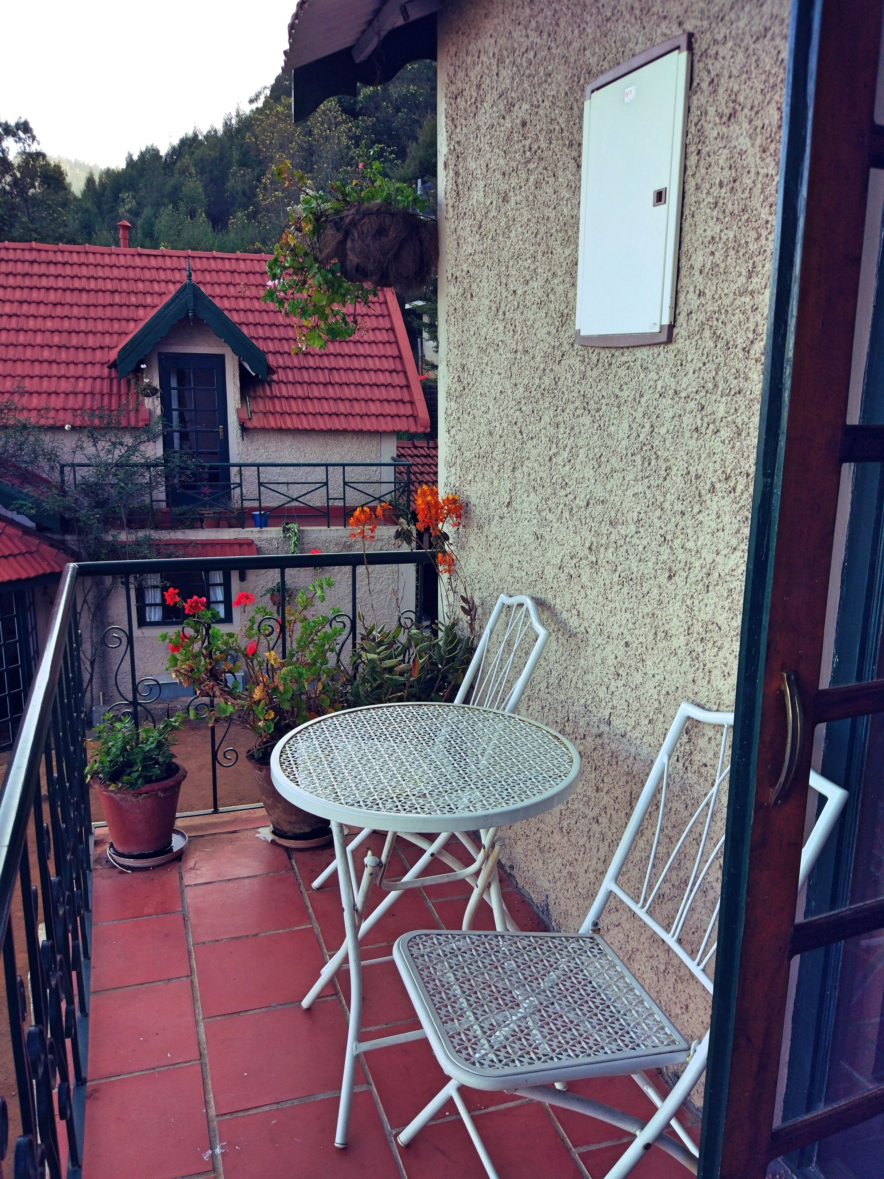Un balcón con una mesa redonda de metal y dos sillas, plantas en macetas y vista a un edificio con tejado rojo.