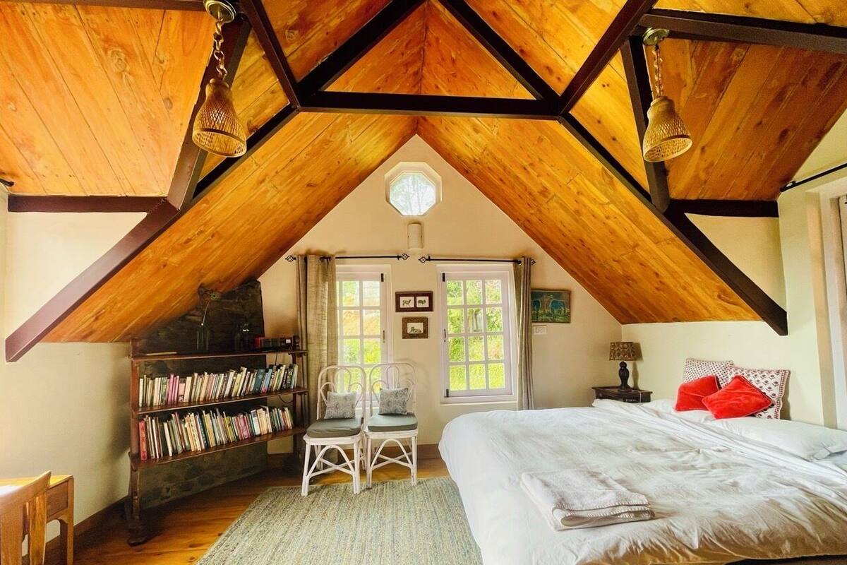 A cozy attic bedroom with a sloped ceiling, a bookshelf, two chairs, a bed with pillows, and a window with curtains.