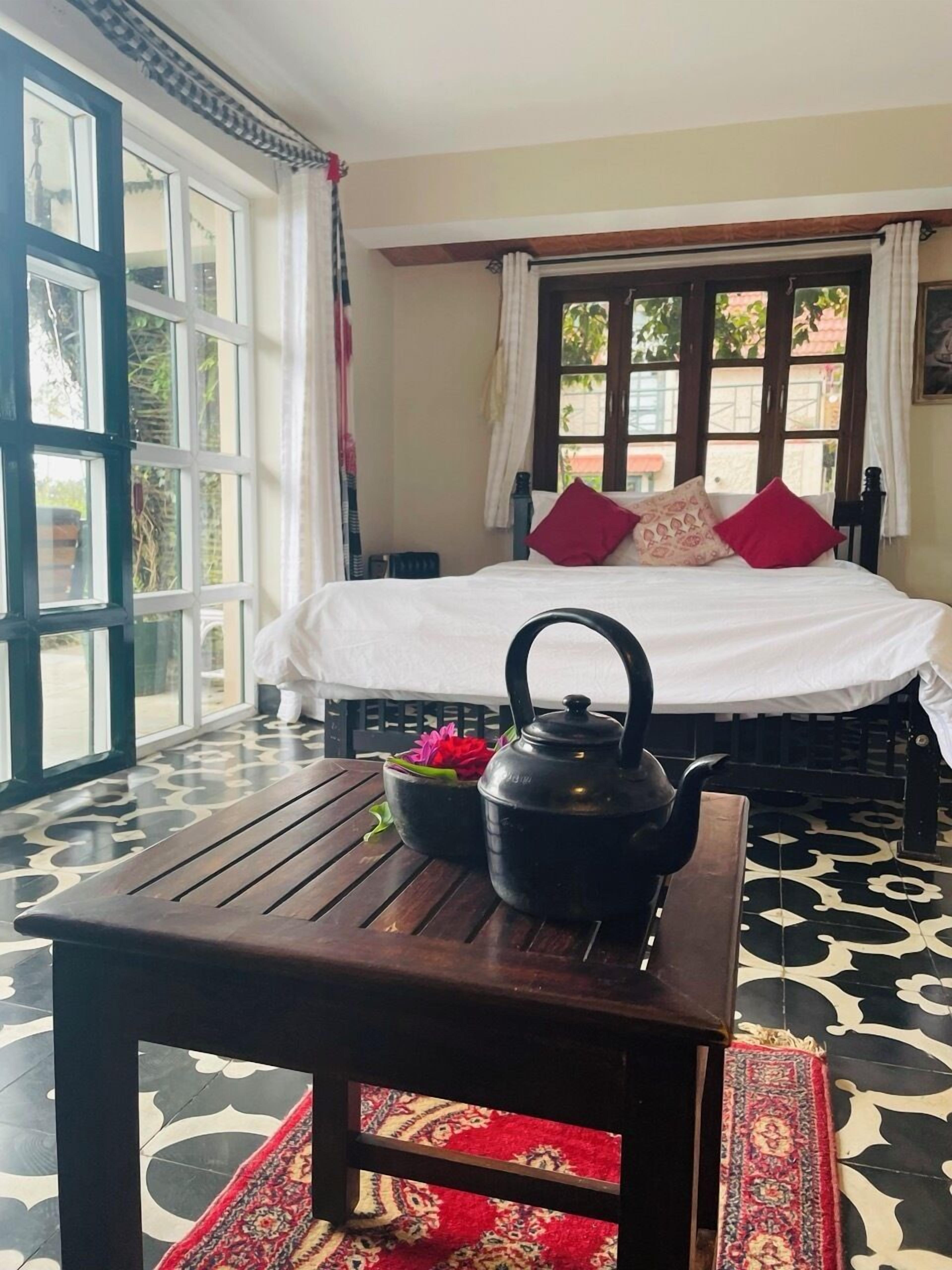 A room with a bed, a table with a teapot and flowers, and a view through large windows.