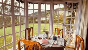 A conservatory with a table set for four, surrounded by wooden chairs, overlooking a grassy landscape. - Stumpfields (Ootacamund)
